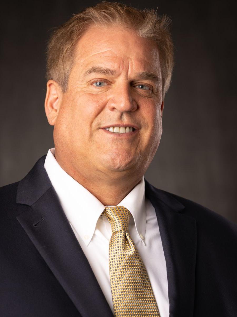 Mr. Jon Dollan, 白人男性 with short grey and brown hair smile wearing a white collared shirt, 带白点的金色领带, and a black suit jacket in front of a grey background.